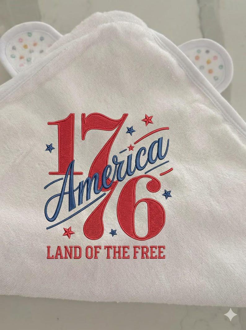 K&ouml;nnte beinhalten: Wei&szlig;es Kapuzenhandtuch f&uuml;r Babys mit gesticktem Text "1776 America" in Rot und Blau, mit Sternen. Der Text "Land of the Free" ist ebenfalls rot gestickt. Das Handtuch hat kleine, wei&szlig;e Ohren mit bunten Punkten.