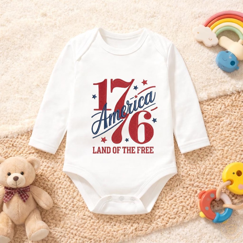 K&ouml;nnte beinhalten: Wei&szlig;er Baby-Body mit langen &Auml;rmeln. Auf der Vorderseite steht der Text "1776 America" in Rot und Blau, mit Sternen und dem Satz "Land of the Free". Ein Teddyb&auml;r und Babyspielzeug befinden sich im Hintergrund.