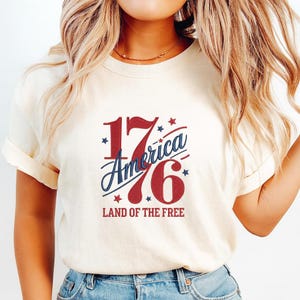 K&ouml;nnte beinhalten: Cremefarbenes T-Shirt mit patriotischem Design. Das Design zeigt die Zahl "1776" in Rot, mit "America" in blauer Schrift. Der Text "LAND OF THE FREE" steht darunter. Sterne umgeben das Design.