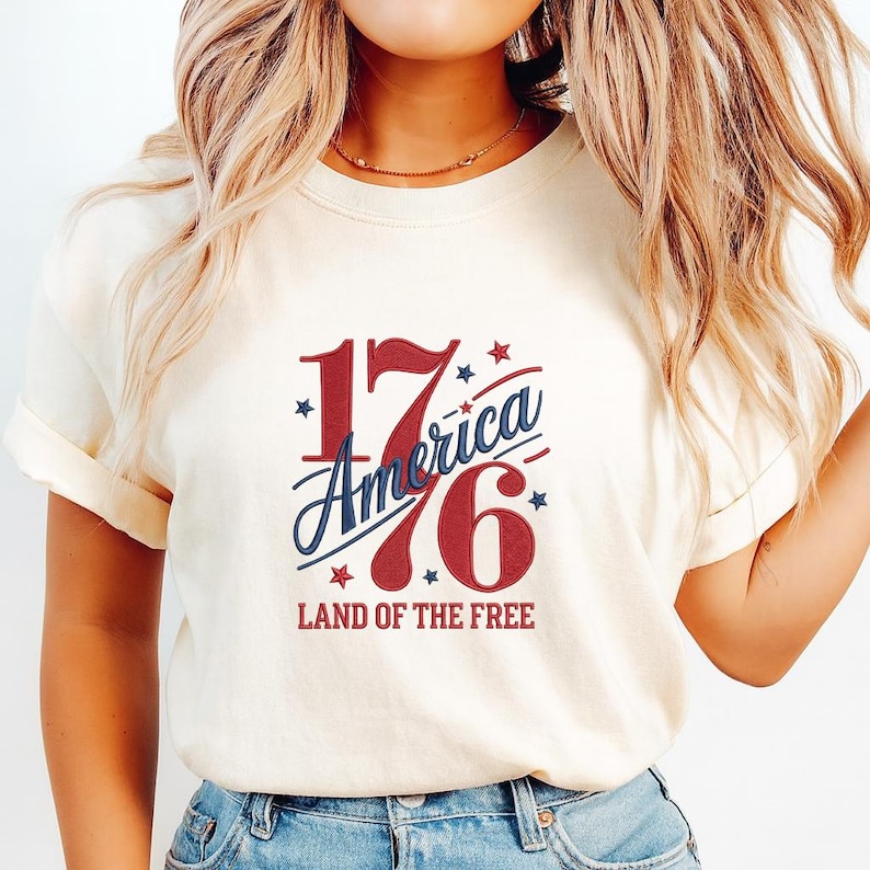K&ouml;nnte beinhalten: Cremefarbenes T-Shirt mit patriotischem Design. Das Design zeigt die Zahl "1776" in Rot, mit "America" in blauer Schrift. Der Text "LAND OF THE FREE" steht darunter. Sterne umgeben das Design.