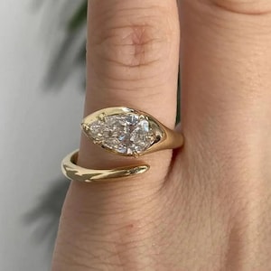 Peut inclure: Une bague ouverte en or avec une pierre précieuse transparente taille marquise montée sur un anneau en or lisse et poli. La bague présente un design ouvert où les deux extrémités ne se rejoignent pas, la pierre étant fixée à une extrémité.