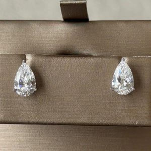 May include: A pair of pear-shaped stud earrings featuring clear, faceted stones set in silver-toned metal. The earrings are displayed on a rectangular, metallic bronze-colored card with a horizontal slot for the earring posts.