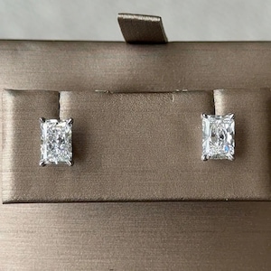 May include: A pair of radiant-cut stud earrings featuring clear, faceted stones set in silver-toned metal prongs. The earrings are displayed on a rectangular, metallic bronze-colored jewelry card with a small tab at the top center.