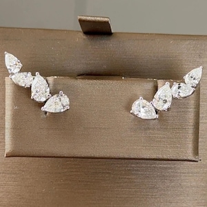 May include: A pair of silver-toned ear climbers featuring a cluster of three clear, pear-shaped stones arranged in a graduated, ascending design. The stones are set in a metallic frame that follows the natural curve of the earlobe for a sleek look.