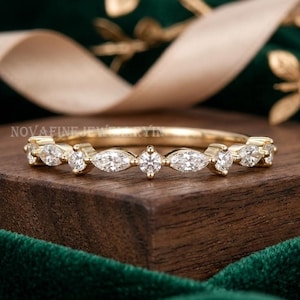 Peut inclure: Une bague en or avec un motif alterné de pierres claires taillées en marquise et en rond sur la moitié supérieure. La bague est placée horizontalement sur un bloc de bois sombre, mettant en valeur un design délicat et symétrique.