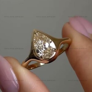 Peut inclure: Une bague en or avec un diamant en forme de poire monté dans un serti clos lisse et poli. L'anneau est en or massif avec un design effilé qui se fond parfaitement dans le sertissage, mettant en valeur la pierre claire et facettée au centre.