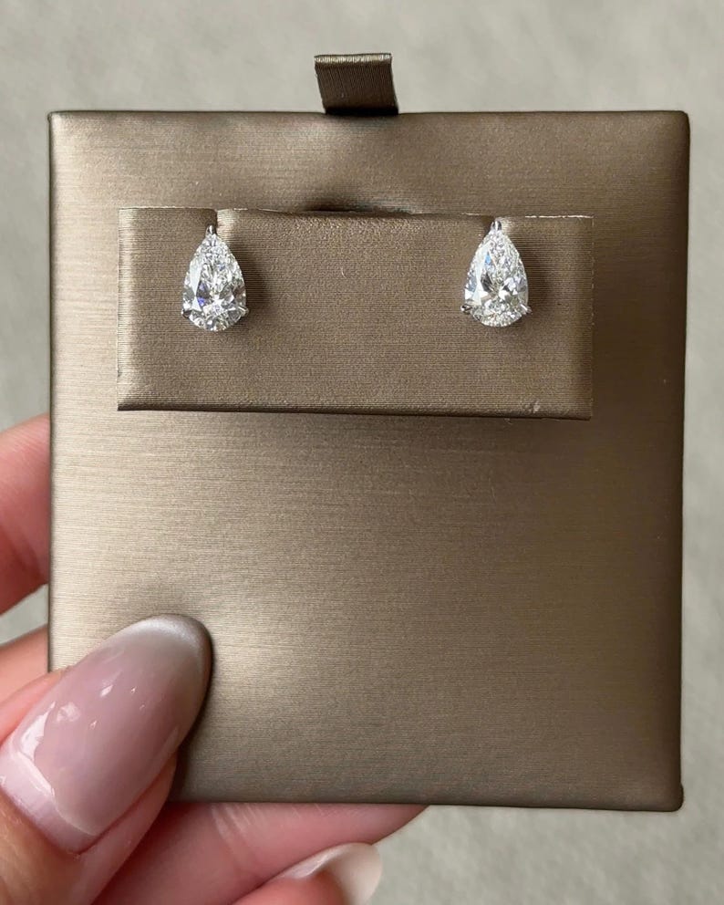 May include: A pair of pear-shaped stud earrings featuring clear, faceted stones set in silver-toned metal. The earrings are displayed on a rectangular, metallic bronze-colored card with a horizontal slot for the earring posts.