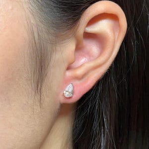 May include: A single pear-shaped stud earring featuring a clear, faceted gemstone set in a metallic prong setting. The earring is positioned on the earlobe, showcasing a brilliant cut design that reflects light with a bright, sparkling appearance.