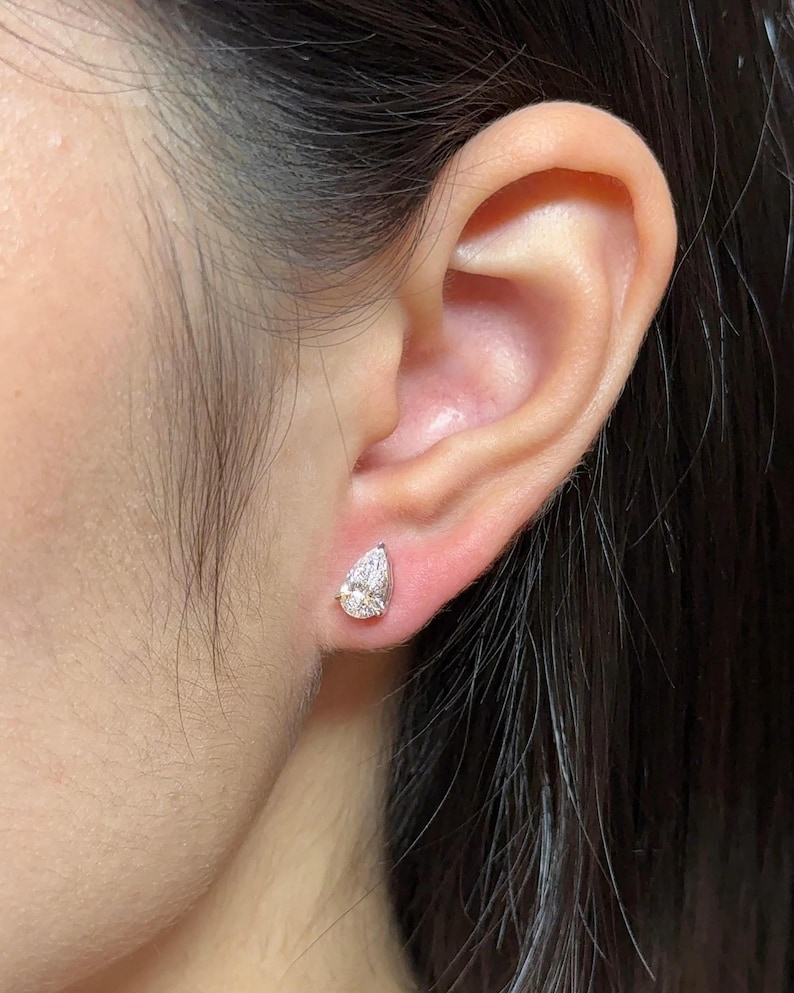 May include: A single pear-shaped stud earring featuring a clear, faceted gemstone set in a metallic prong setting. The earring is positioned on the earlobe, showcasing a brilliant cut design that reflects light with a bright, sparkling appearance.