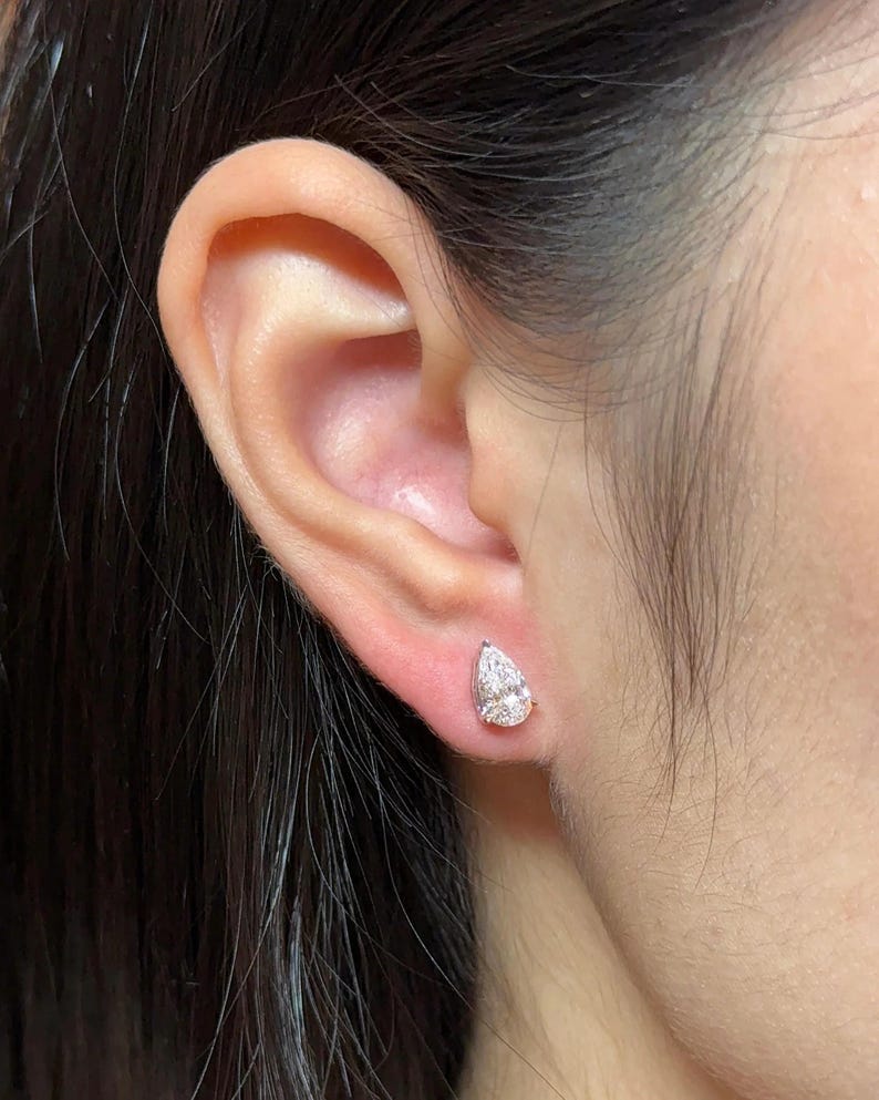 May include: A single pear-shaped stud earring featuring a clear, faceted gemstone set in a polished silver-toned metal base. The earring is positioned securely in the earlobe, showcasing a brilliant, reflective surface and a classic teardrop silhouette.