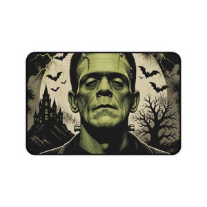 Puede incluir: Una alfombrilla de escritorio rectangular con una ilustración en tonos verdes del monstruo de Frankenstein centrada frente a una gran luna llena. El fondo incluye la silueta oscura de un castillo, un árbol desnudo, murciélagos volando y rayos.