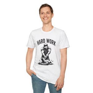 Può includere: Una t-shirt bianca a maniche corte con una stampa grafica nera che raffigura una persona con un berretto che usa una mazza per rompere delle pietre. Il testo "HARD WORK" è stampato in un carattere nero, audace e arcuato.