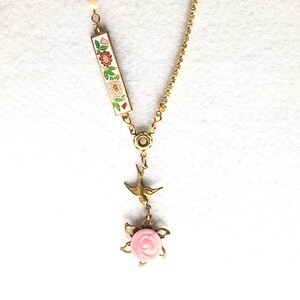May include: A gold chain necklace with a pink rose and a bird pendant. The pendant is attached to a rectangular charm with a floral design.