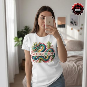 May include: White t-shirt featuring a colorful dragon design with the word "MAHJONG" in block letters. The dragon is teal, blue, and yellow, surrounded by bamboo and pink flowers. The shirt is being worn by a person.
