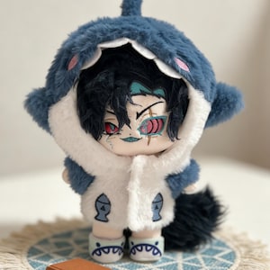 May include: A plush doll wearing a blue shark hoodie with a white and blue jacket. The doll has black hair and red eyes. It is standing on a small, round, blue and white patterned mat with a small brown book.