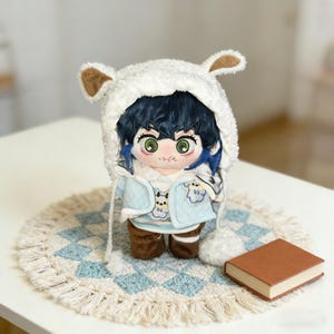 Puede incluir: Muñeco de peluche con pelo azul y ojos verdes, que lleva un gorro blanco con orejas de oso y una chaqueta azul claro con parches de oso. El muñeco está sobre una alfombra con estampado azul y blanco, con un libro pequeño cerca.