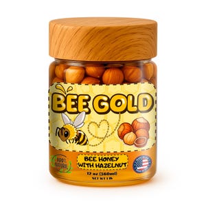 Puede incluir: Un tarro de cristal de miel Bee Gold con tapa de madera. La etiqueta presenta una abeja de dibujos animados, avellanas y el texto "BEE GOLD", "BEE HONEY WITH HAZELNUT" y "12 oz (360ml) NET WT 1 lb". El tarro está lleno de avellanas.