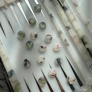 May include: A backgammon set with a white playing surface and clear, round game pieces. The pieces are decorated with small flowers and leaves. The board has brown and black points and a clear lid.
