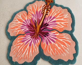 May include: A tufted rug shaped like a hibiscus flower, featuring large petals in shades of peach and orange with white vein-like details. The centre is a deep magenta and dark red, with a prominent yellow stamen extending upwards, all bordered by a thick teal edge.