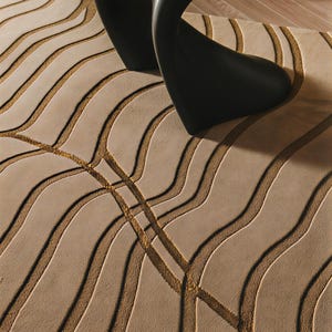 May include: A beige area rug featuring a textured pattern of raised, undulating wave lines in a darker tan shade, with a smooth black curved furniture piece resting on the surface of the rug, partially obscuring the view of the floor covering.