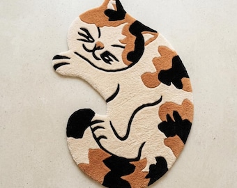 May include: A tufted rug shaped like a curled-up calico cat. The rug features a cream-coloured base with patches of black and orange-brown fur. The cat has a stylised face with closed eyes and a small mouth, with black outlines defining its form.