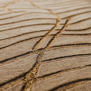 May include: Close-up of a beige rug with a textured, ridged surface. The rug features a golden, linear design that meanders across the surface, creating a unique pattern. The rug's design adds a touch of elegance.