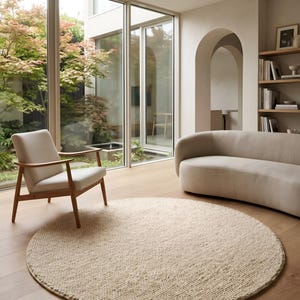May include: A round, cream-coloured, textured wool rug with a thick, looped pile sits on a light-toned wooden floor. The rug is positioned in a room featuring a light-coloured armchair with wooden arms and a curved, light-coloured sofa.