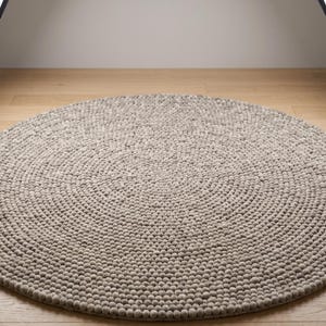 Könnte beinhalten: Ein runder Teppich, der aus zahlreichen kleinen, dicht aneinandergereihten gefilzten Wollkugeln in einer neutralen Farbpalette aus Hellgrau und Beige besteht. Die einzelnen Kugeln sind in konzentrischen Kreisen angeordnet.