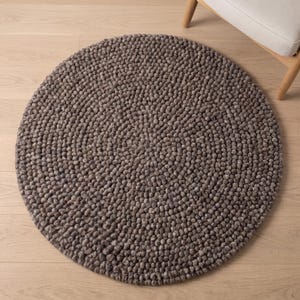 Puede incluir: Una alfombra circular construida con numerosas pequeñas bolas de lana afieltrada de color gris oscuro dispuestas en un patrón denso y uniforme. La alfombra presenta una superficie texturizada y abultada creada por los elementos esféricos.