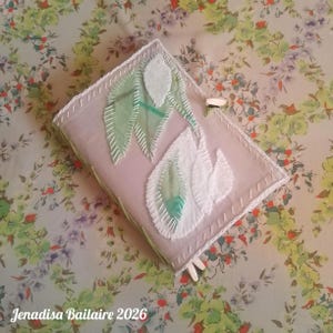 May include: A rectangular fabric-covered journal featuring a light pink cover with white and green leaf-shaped fabric appliqués. The edges are finished with white decorative stitching, and a small wooden toggle closure is visible on the side.