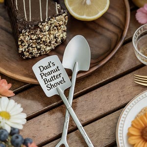 Peut inclure: Deux pelles à beurre de cacahuète argentées sont croisées sur une table en bois. L'une des pelles porte l'inscription "Dad's Peanut Butter Shovel". Une part de gâteau au chocolat aux noix et une moitié de citron sont à proximité.