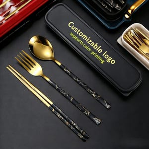 Puede incluir: Un juego de cubiertos negro y dorado con una cuchara, un tenedor y palillos. Los utensilios tienen cabezas doradas y mangos negros con diseños geométricos dorados. El juego incluye un estuche negro con el texto "Customizable logo supports color printing."