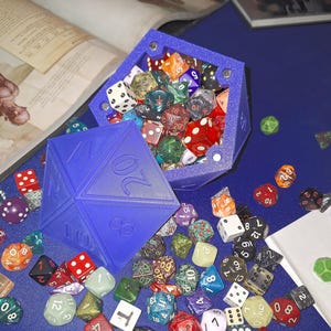 May include: A collection of colorful polyhedral dice scattered on a blue surface, with a blue D20 die and a blue container filled with dice. The dice are in various colors and styles, including red, white, green, and clear.