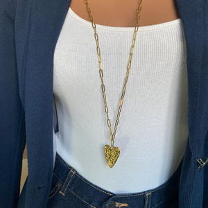 May include: A gold-colored necklace with a textured heart pendant. The long chain is made of oval links. The necklace is worn over a white tank top and dark blue denim jeans. A navy blue blazer is worn over the tank top.