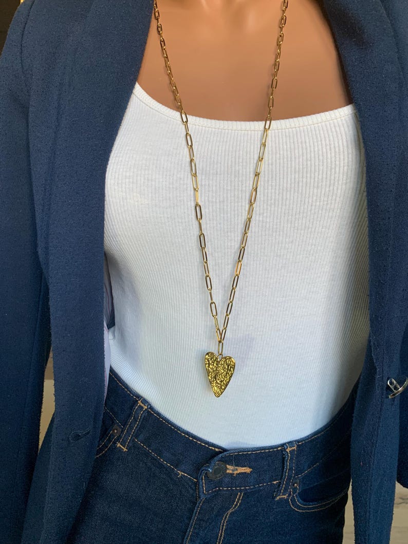 May include: A gold-colored necklace with a textured heart pendant. The long chain is made of oval links. The necklace is worn over a white tank top and dark blue denim jeans. A navy blue blazer is worn over the tank top.