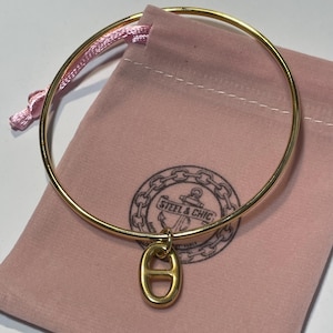 Pode incluir: Uma pulseira de metal dourado com um pingente oval que possui uma barra horizontal no centro. A pulseira está posicionada sobre uma bolsa de tecido rosa claro que exibe um logotipo circular com uma âncora e o texto STEEL & CHIC.