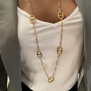 Peut inclure: Un long collier doré composé d'une série de maillons rectangulaires allongés entrecoupés de quatre maillons ovales, chacun doté d'une barre horizontale au centre, créant un motif géométrique structuré et répétitif.