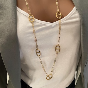 Peut inclure: Un long collier doré avec des maillons ovales et un motif de chaîne. Le collier est porté sur un haut blanc et un blazer gris. Le design du collier est simple et élégant.