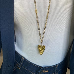 May include: A gold-colored necklace with a textured heart pendant. The long chain is made of rectangular links. The necklace is worn over a white ribbed tank top and dark blue denim jeans.