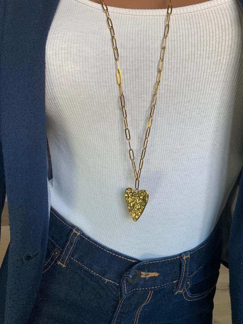 May include: A gold-colored necklace with a textured heart pendant. The long chain is made of rectangular links. The necklace is worn over a white ribbed tank top and dark blue denim jeans.