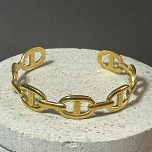 Peut inclure: Un bracelet manchette doré présentant un motif de maillons de chaîne répétitifs. Chaque maillon est de forme ovale avec une barre horizontale centrale, créant un motif cohérent sur tout le bracelet ouvert. Le métal a une finition polie.