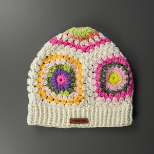 Può includere: Un berretto all'uncinetto fatto a mano con un motivo a granny square. Il cappello presenta una base color crema con quadrati colorati nelle tonalità di rosa, giallo, verde e viola. Una piccola etichetta marrone con testo è cucita sull'orlo.