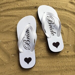 May include: A pair of white flip-flops featuring black text that reads Bride in a cursive script and a solid black heart icon printed on the footbed. The sandals have white rubber straps and a black sole, designed for casual footwear use.