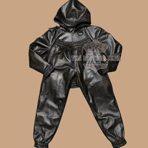May include: Black leather hooded jumpsuit with a drawstring hood, elastic cuffs, and a zippered chest pocket. The jumpsuit has a relaxed fit with elastic at the waist and ankles. The text "THE LEATHER SKIN" is visible on the chest.