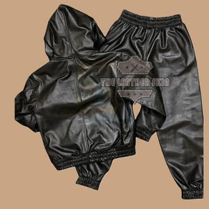 May include: Black leather hooded jacket and matching trousers set. The jacket features a hood, zip closure, and elasticated cuffs and hem. The trousers have an elasticated waistband and cuffs. The text "THE LEATHER SKIN" is visible on the back of the jacket.