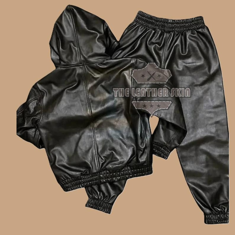 May include: Black leather hooded jacket and matching trousers set. The jacket features a hood, zip closure, and elasticated cuffs and hem. The trousers have an elasticated waistband and cuffs. The text "THE LEATHER SKIN" is visible on the back of the jacket.