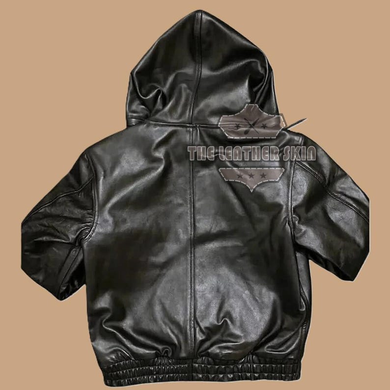 May include: Rear view of a black leather hooded jacket. The jacket features a zip closure, a hood, and a ribbed hem. The text "THE-LEATHER SKIN" is displayed on the back of the jacket.