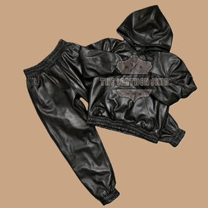 May include: Black leather hoodie and matching trousers set. The hoodie features a drawstring hood, elasticated waist and cuffs. The trousers have an elasticated waistband and cuffs. The text "THE LEATHER SKIN" is visible on the hoodie.