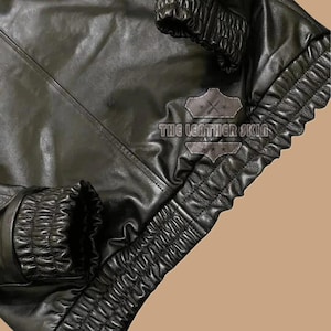May include: Close-up of a black leather jacket with gathered detailing on the cuffs and lower edge. The jacket has a smooth, shiny finish and the text "THE LEATHER SKIN" is visible.