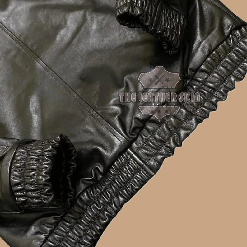 May include: Close-up of a black leather jacket with gathered detailing on the cuffs and lower edge. The jacket has a smooth, shiny finish and the text "THE LEATHER SKIN" is visible.